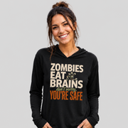 A woman wearing a black long-sleeve hoodie with a graphic print that reads 'ZOMBIES EAT BRAINS don't worry YOU'RE SAFE' in beige and orange text, paired with blue jeans, against a solid gray background.