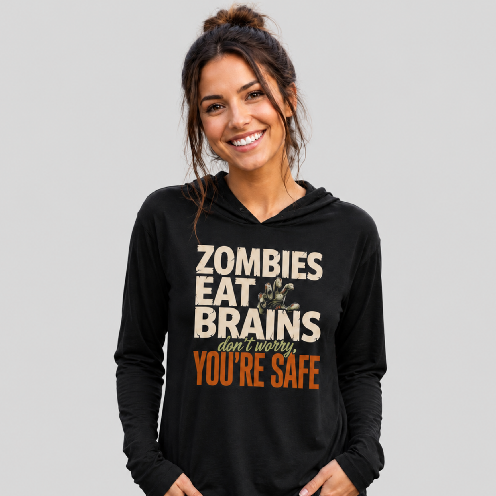 A woman wearing a black long-sleeve hoodie with a graphic print that reads 'ZOMBIES EAT BRAINS don't worry YOU'RE SAFE' in beige and orange text, paired with blue jeans, against a solid gray background.