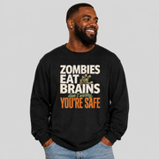 A smiling man wearing a black long-sleeve graphic t-shirt with the text 'ZOMBIES EAT BRAINS don't worry YOU'RE SAFE' in white, green, and orange, paired with light blue jeans, against a solid gray background.